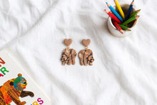 Load image into Gallery viewer, Marbled brown bear earrings with brown heart tops beside a storybook and coloured pencils
